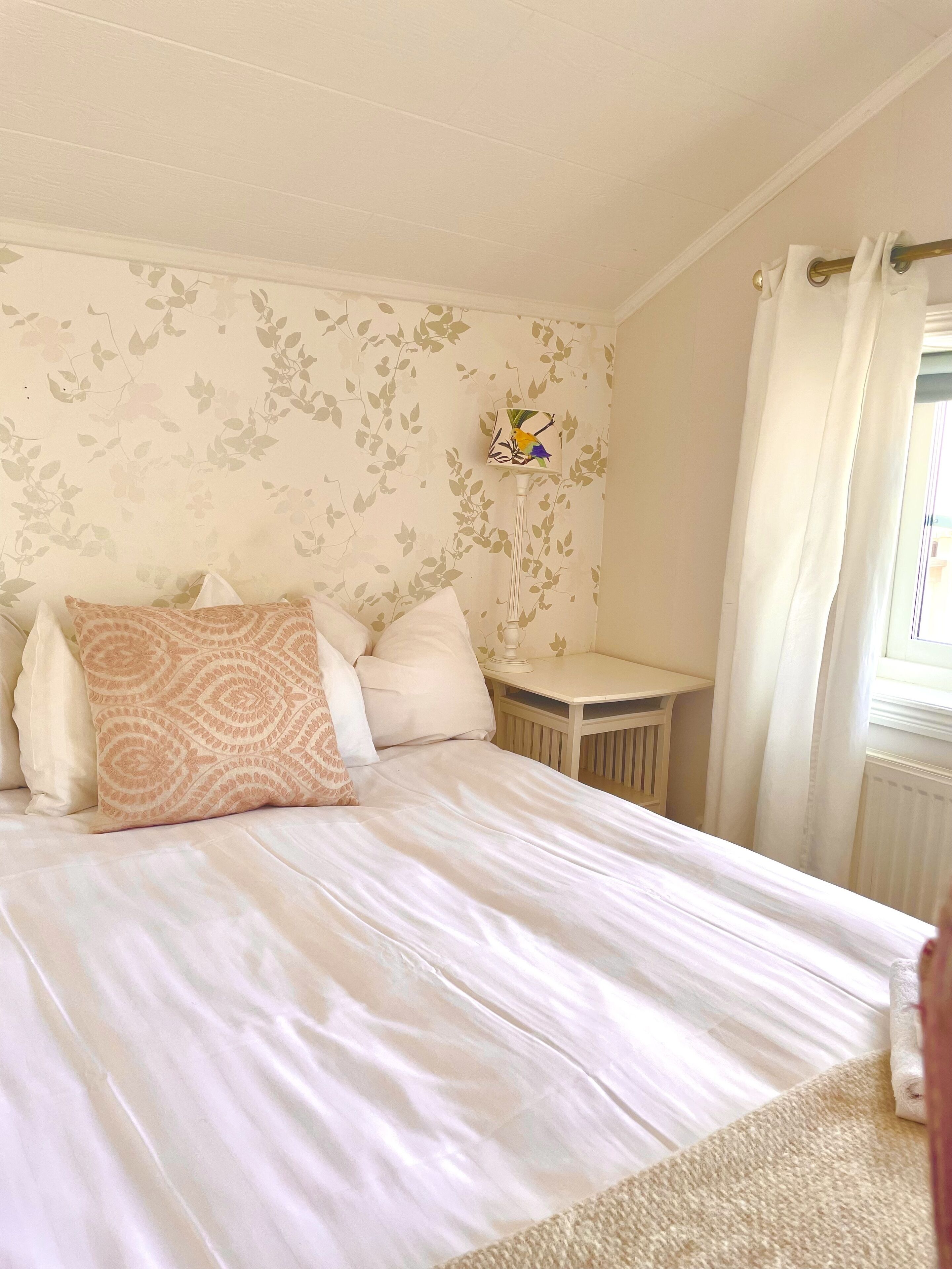 Een net opgemaakt bed met witte lakens en gestreepte kussens, een klein nachtkastje met een lamp, en een raam met gordijnen.