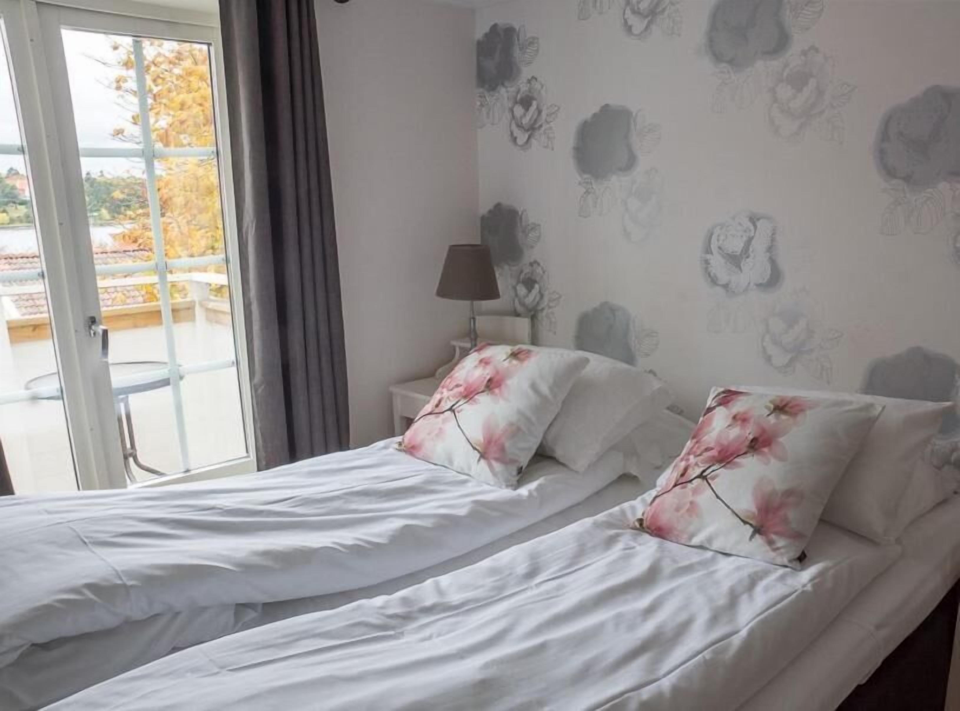 Ein Schlafzimmer mit zwei Betten, einem Fenster mit Vorhängen, einer Lampe auf einem Nachttisch und blumigem Tapetenmuster.
