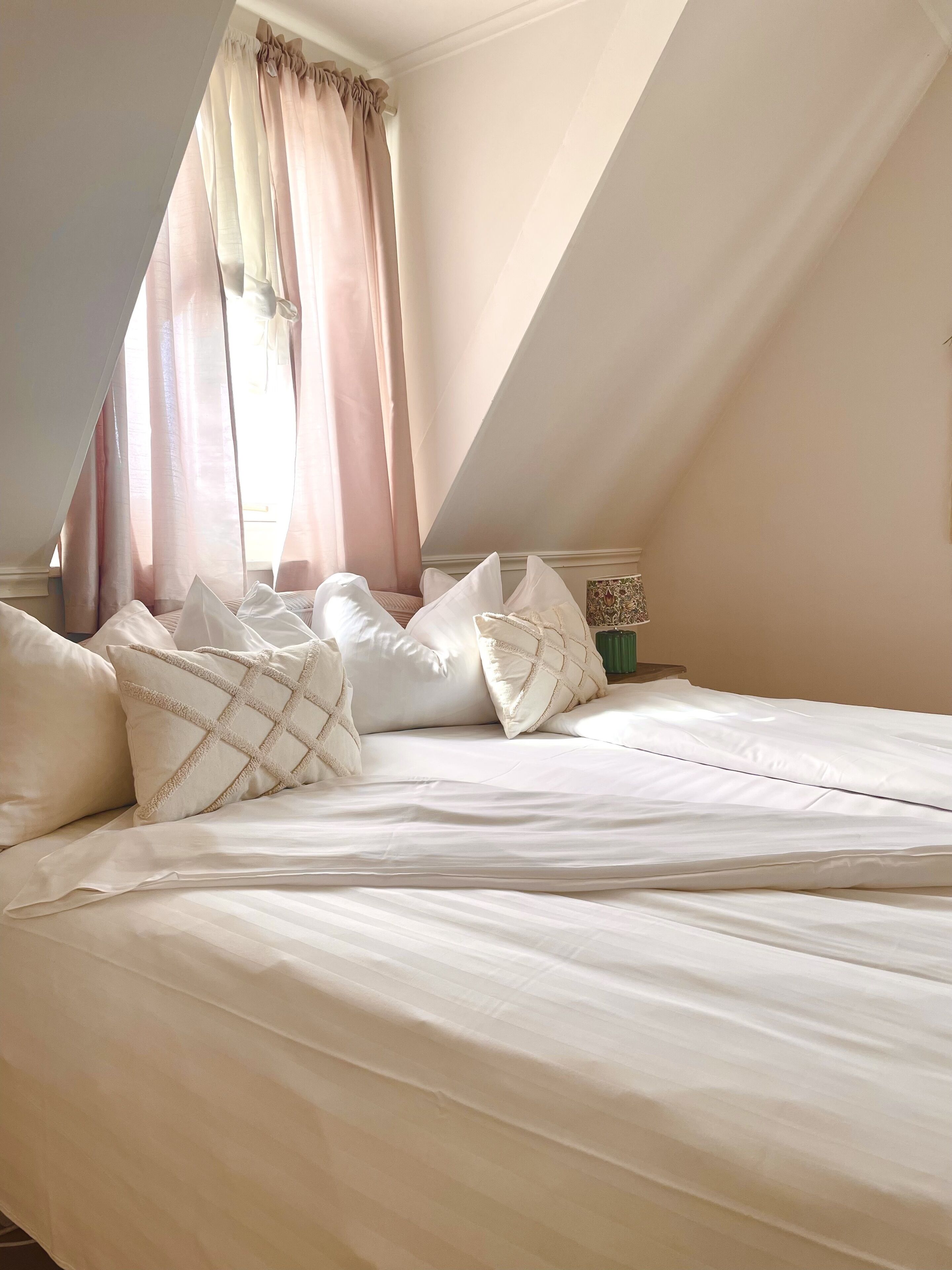 Un dormitorio con techo inclinado, cama con ropa blanca, varios almohadones y cortinas traslúcidas.