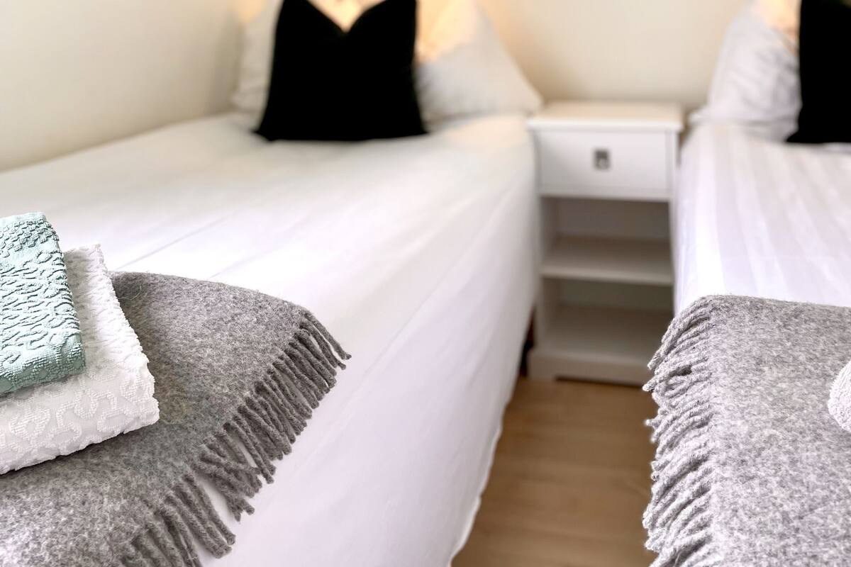Une chambre d’hôtel avec deux lits, chacun agrémenté de draps blancs et d’oreillers noirs. On y trouve des tables de chevet avec des lampes, un tableau au mur et un plaid gris sur l’un des lits.
