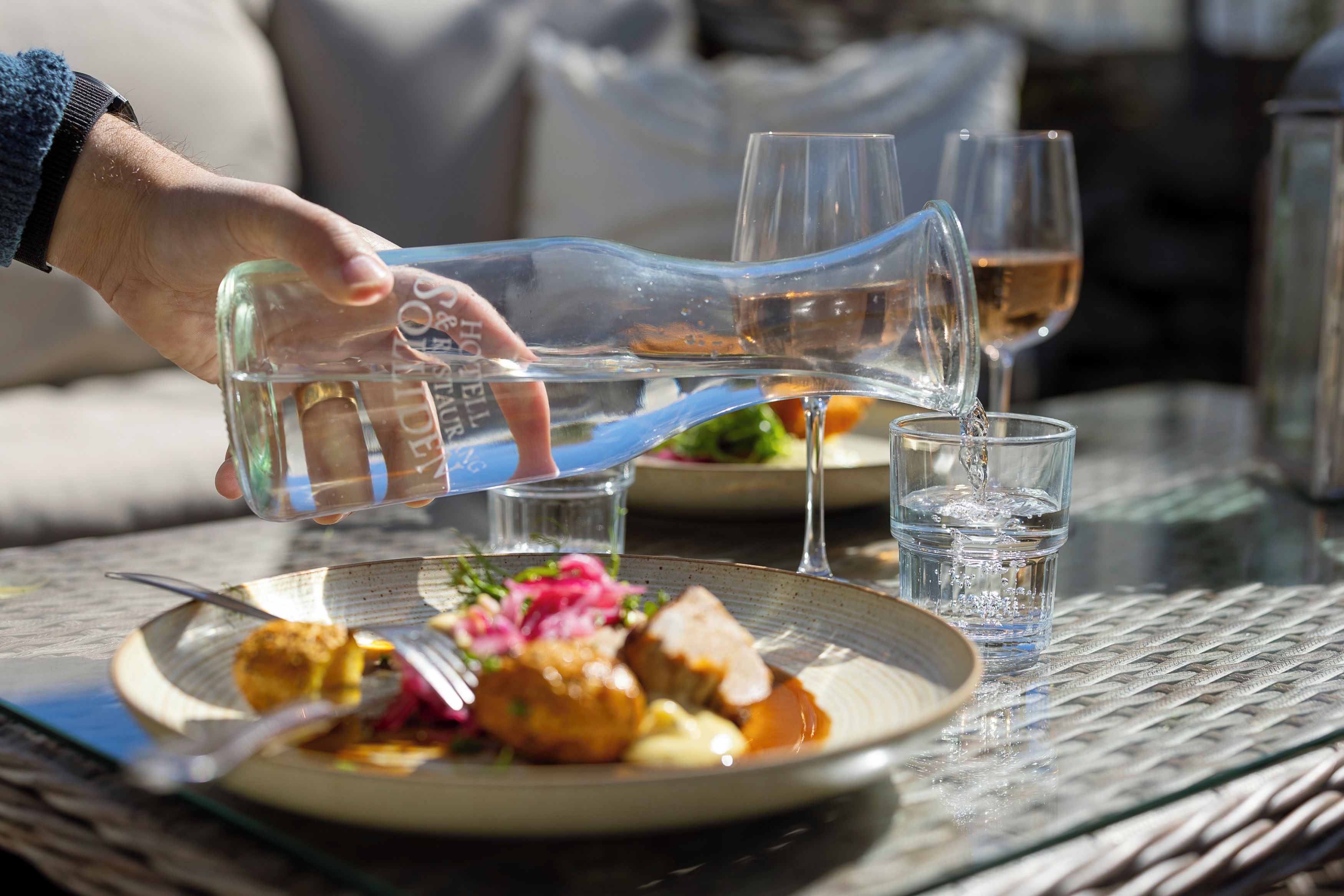 Une personne verse de l’eau dans un verre posé sur une table, à côté d’une assiette de nourriture et d’un verre à vin.