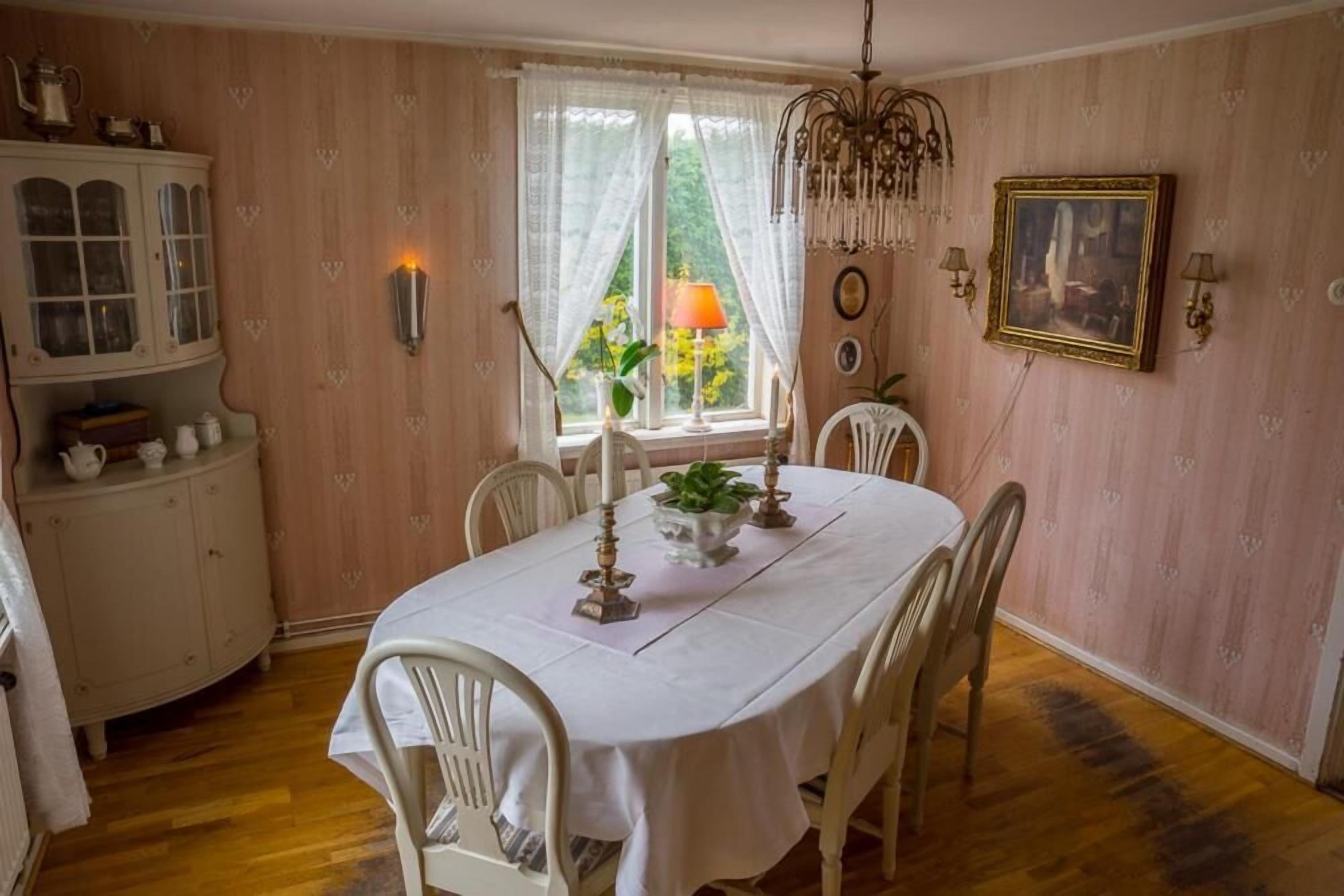 Comedor con una mesa blanca dispuesta para cuatro personas, rodeada de sillas blancas. A la izquierda, un aparador con puertas de cristal; arriba, un candelabro y, al fondo, una ventana con cortinas.