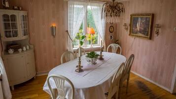 Comedor con una mesa blanca dispuesta para cuatro personas, rodeada de sillas blancas. A la izquierda, un aparador con puertas de cristal; arriba, un candelabro y, al fondo, una ventana con cortinas.