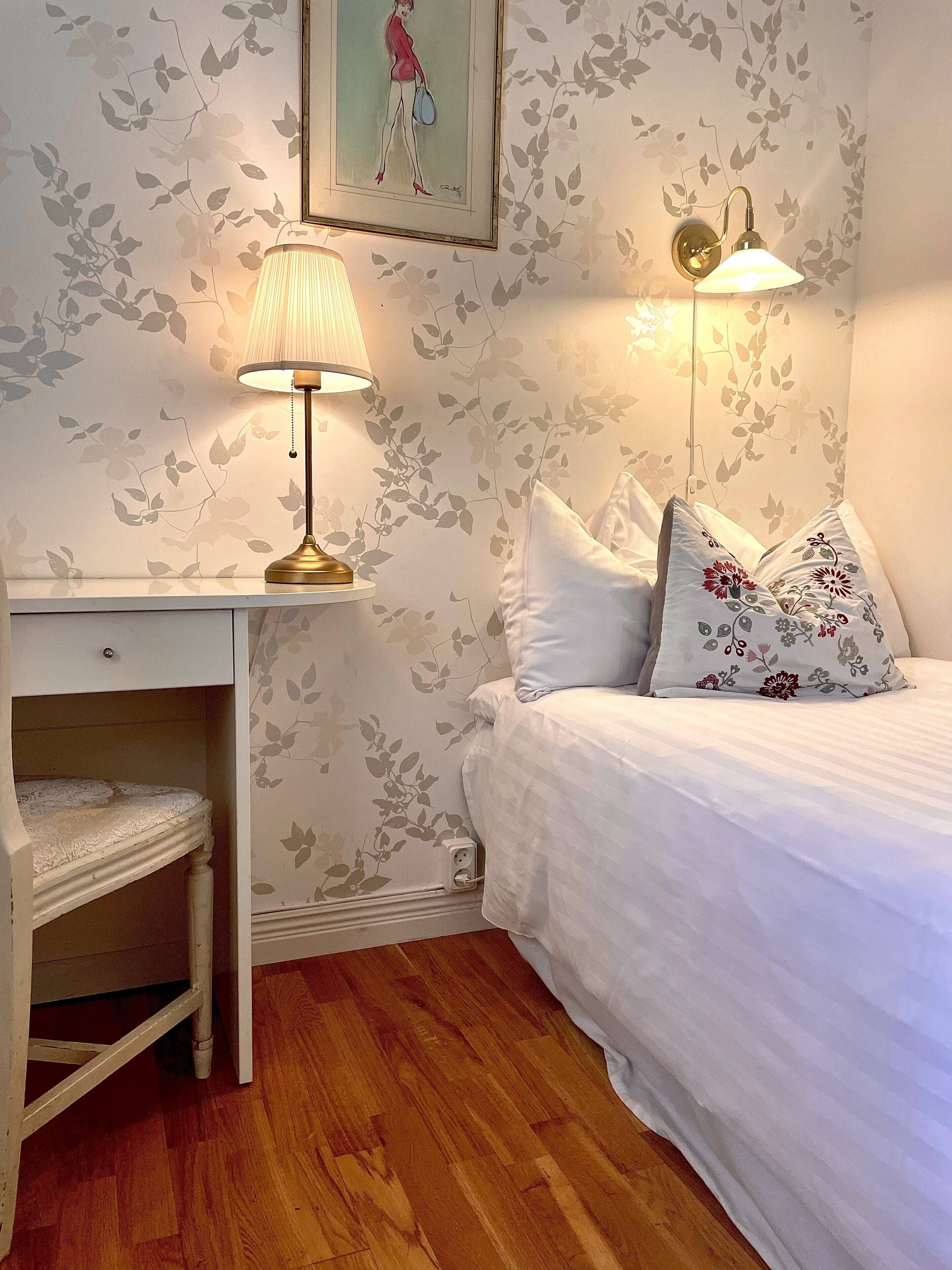 A bedroom with a bed, bedside table, lamp, and a small desk with a chair.