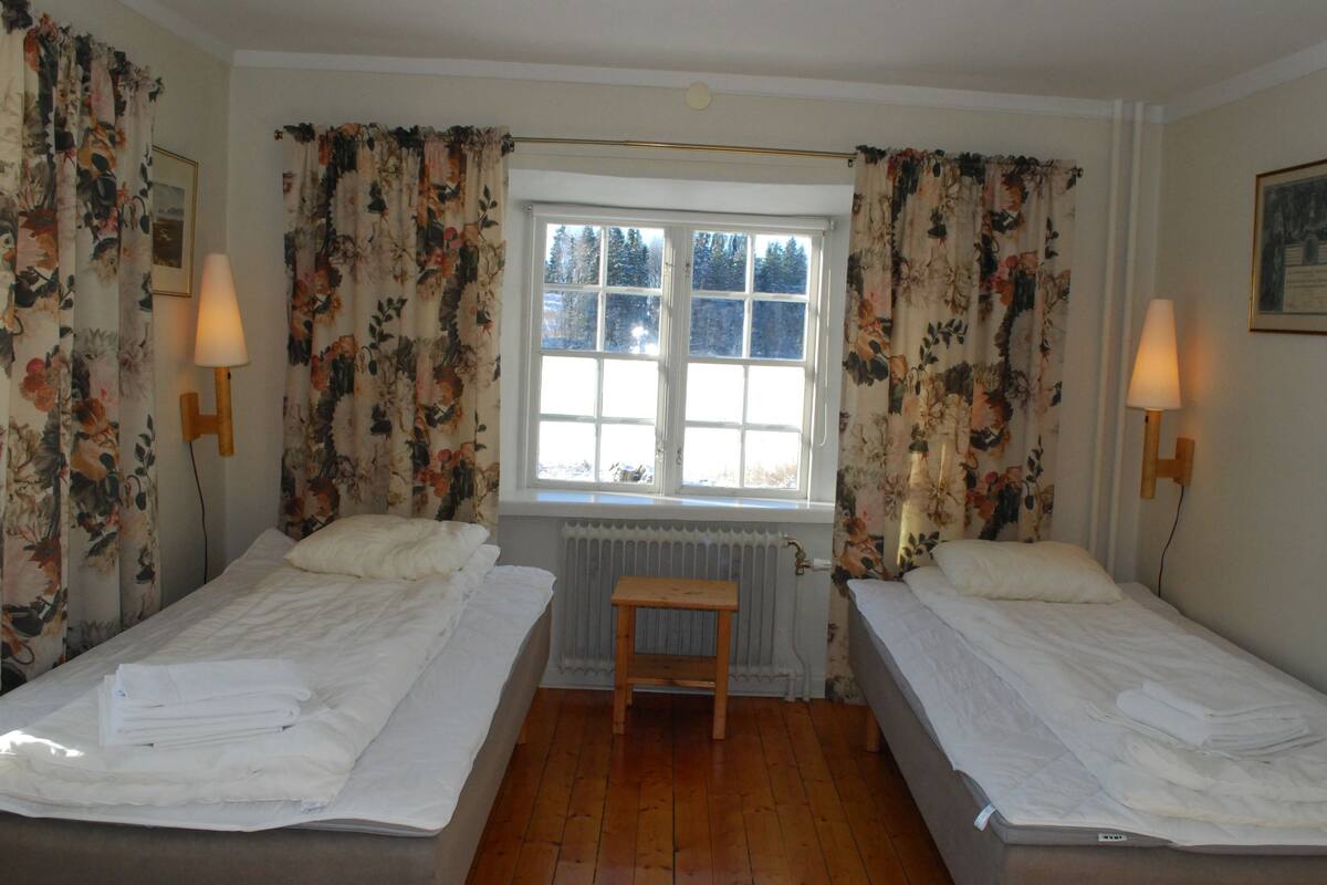 A room with two beds, a wooden bench, and a window with curtains.