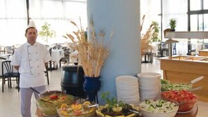 A buffet setup with various dishes, a chef in uniform, and a large pillar with decorative elements. - Nina Beach Hotel (Kos)