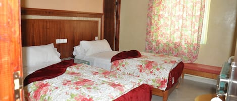 A hotel room with two beds, a wooden headboard, a bench, a window with patterned curtains, and a small table with a bottle.
