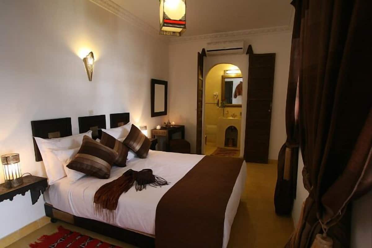 Una habitación de hotel con una cama grande, una mesita de noche con una lámpara, un espejo y una puerta que da a un baño.