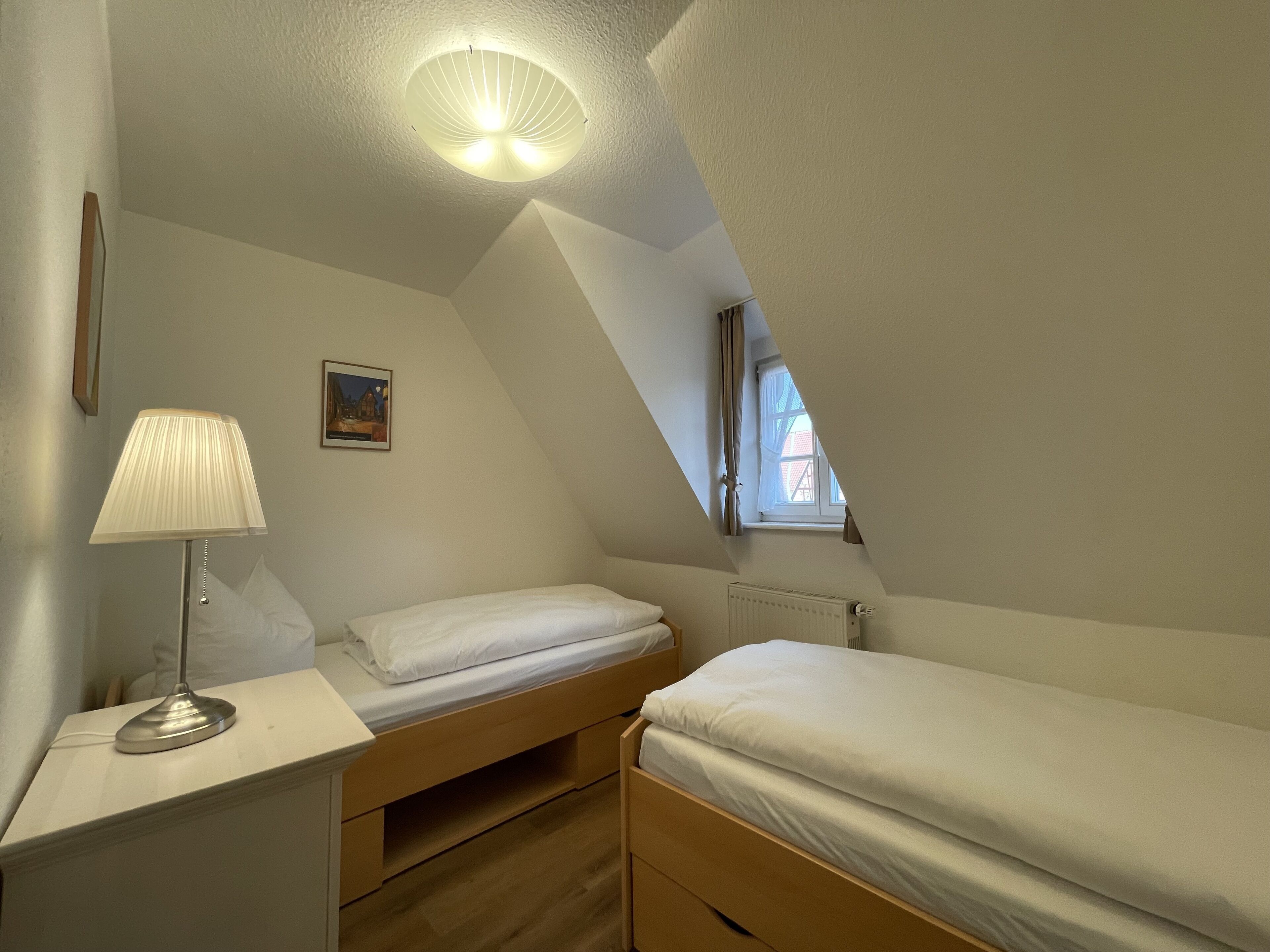 A small, well-lit room with two single beds, a bedside table with a lamp, a radiator, and a painting on the wall.