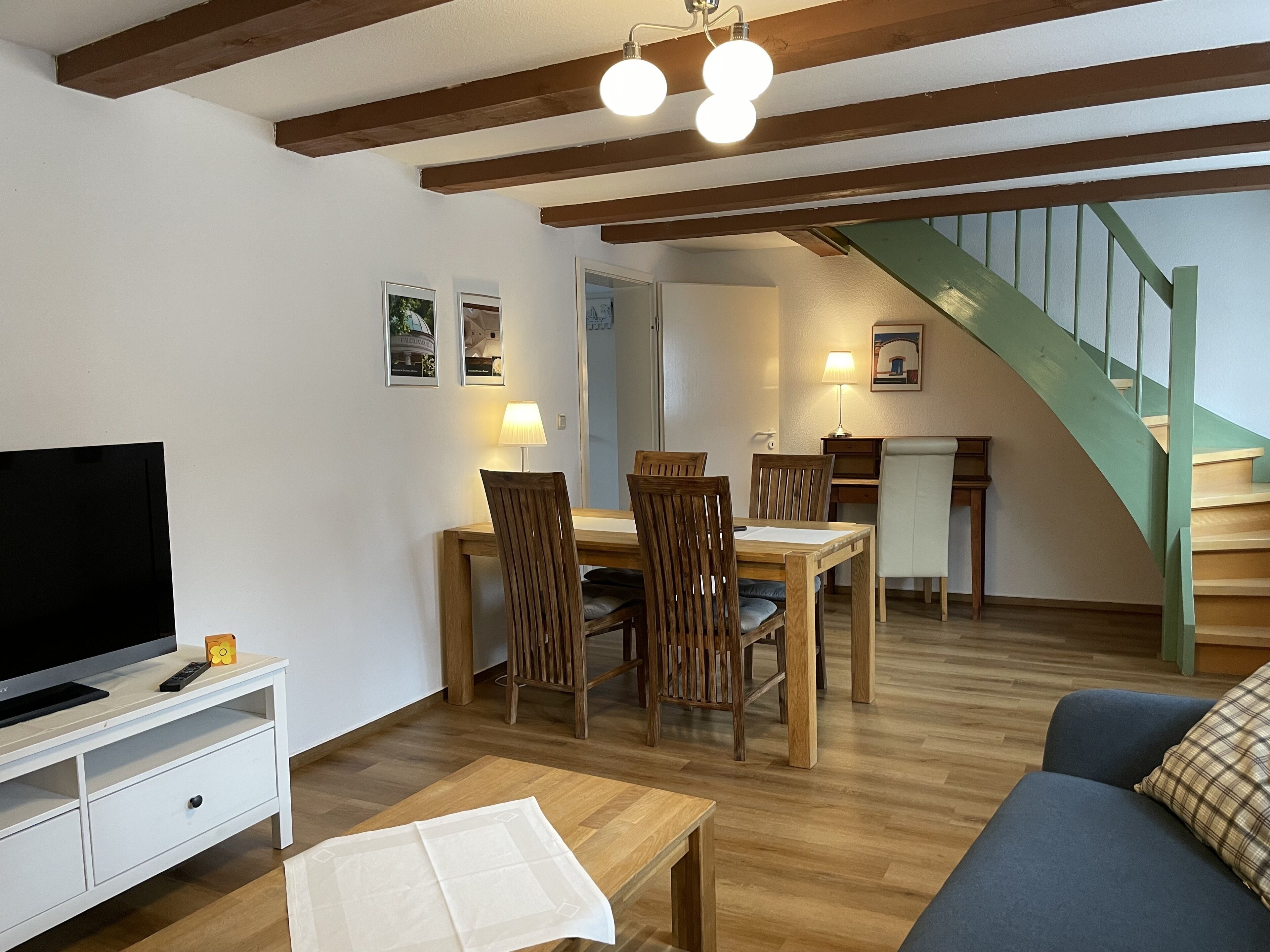 A living room with a TV, wooden dining table, chairs, a sofa, and a staircase.