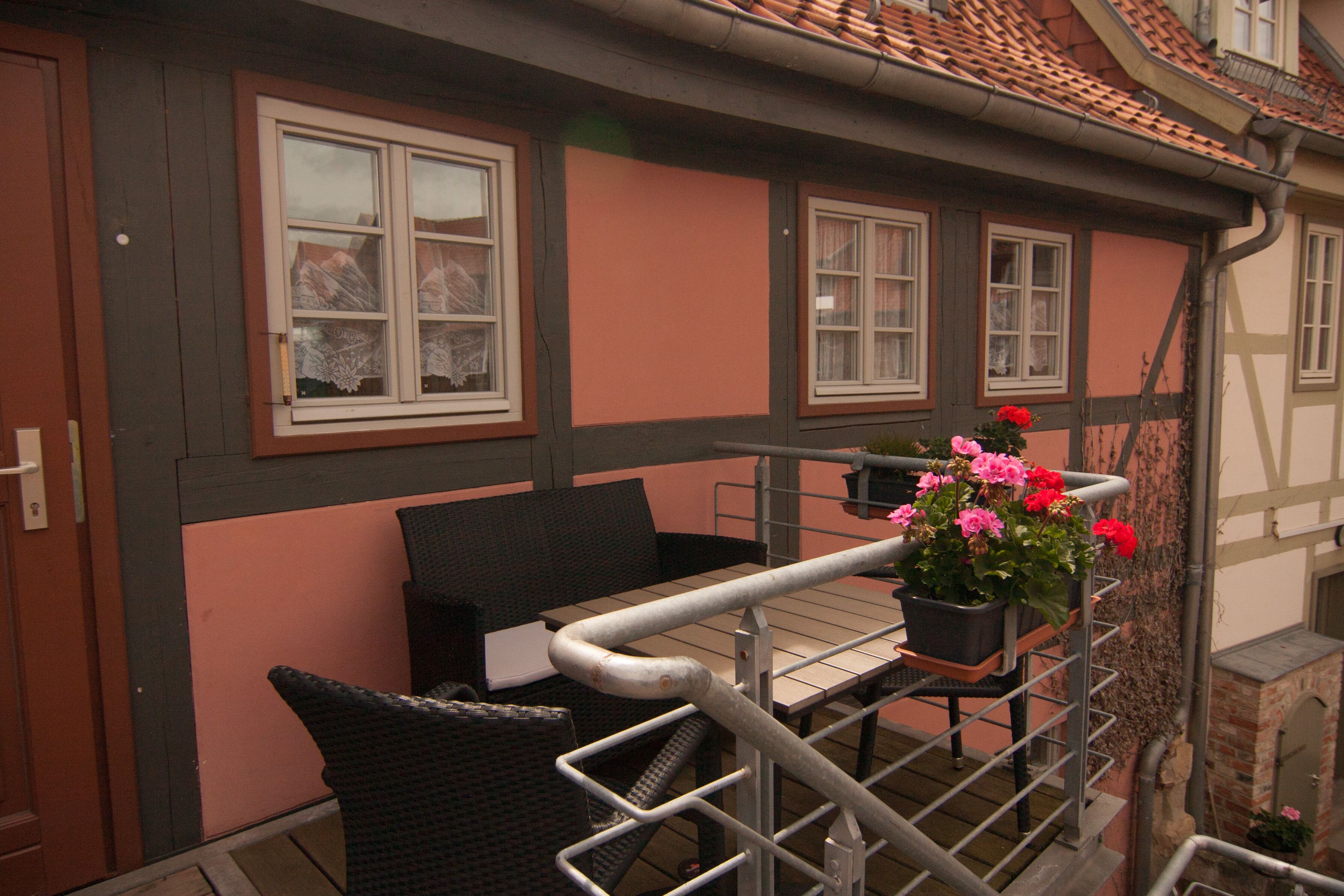 Ein Balkon mit Korbmöbeln, einem Blumentopf mit Blumen und Blick auf ein Gebäude mit Ziegeldach.