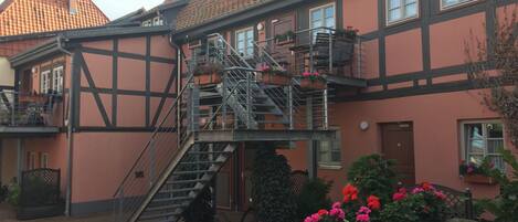 A traditional European building with a staircase leading to an upper floor, surrounded by potted plants and flowers.