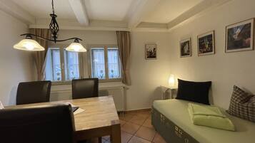 Une pièce chaleureuse avec une table à manger, des chaises, un canapé avec des coussins, une table d’appoint avec une lampe, et des tableaux encadrés accrochés au mur.