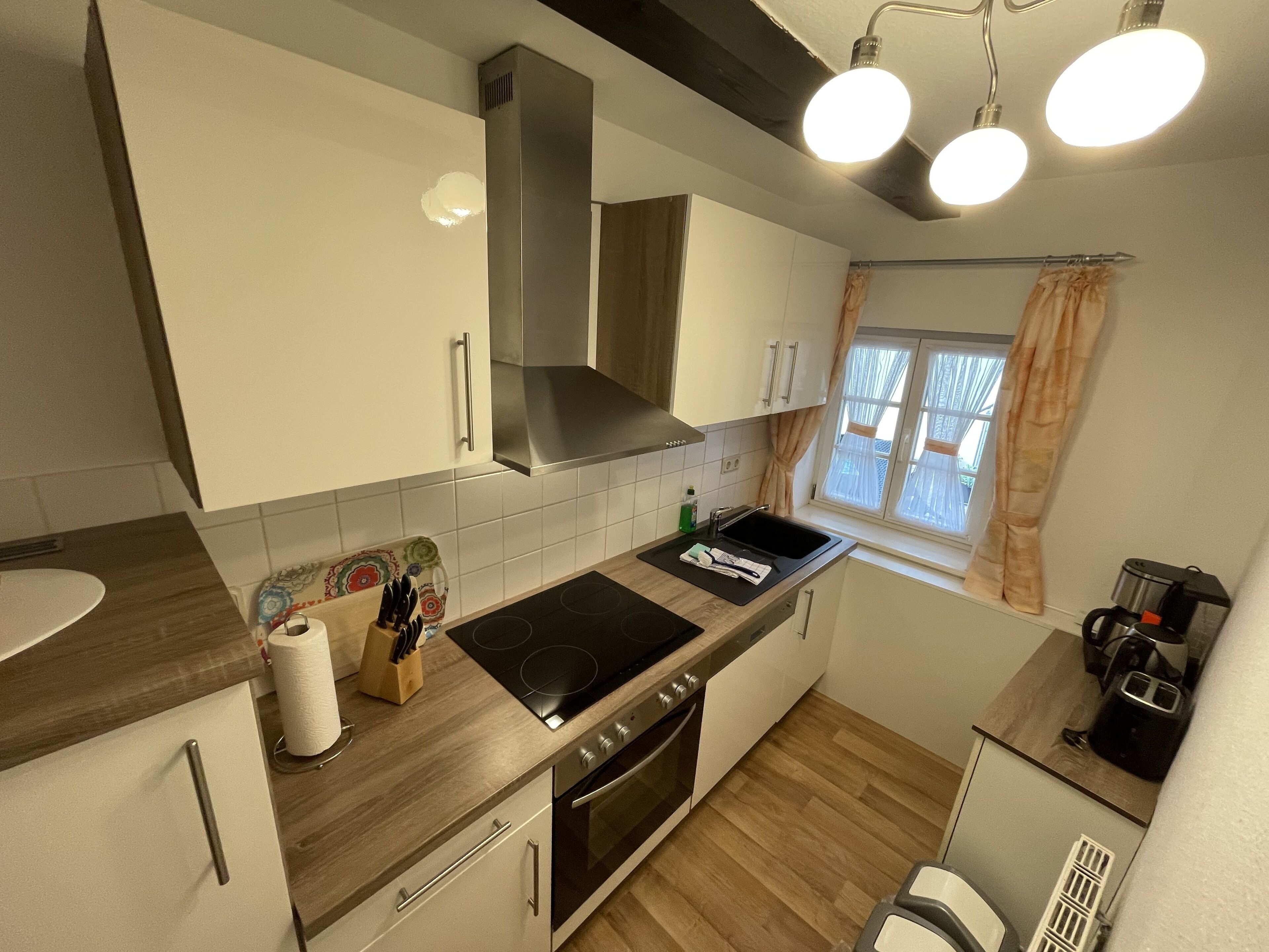 A modern kitchen with white cabinets, a stainless steel range hood, a black stovetop, and a window with curtains.