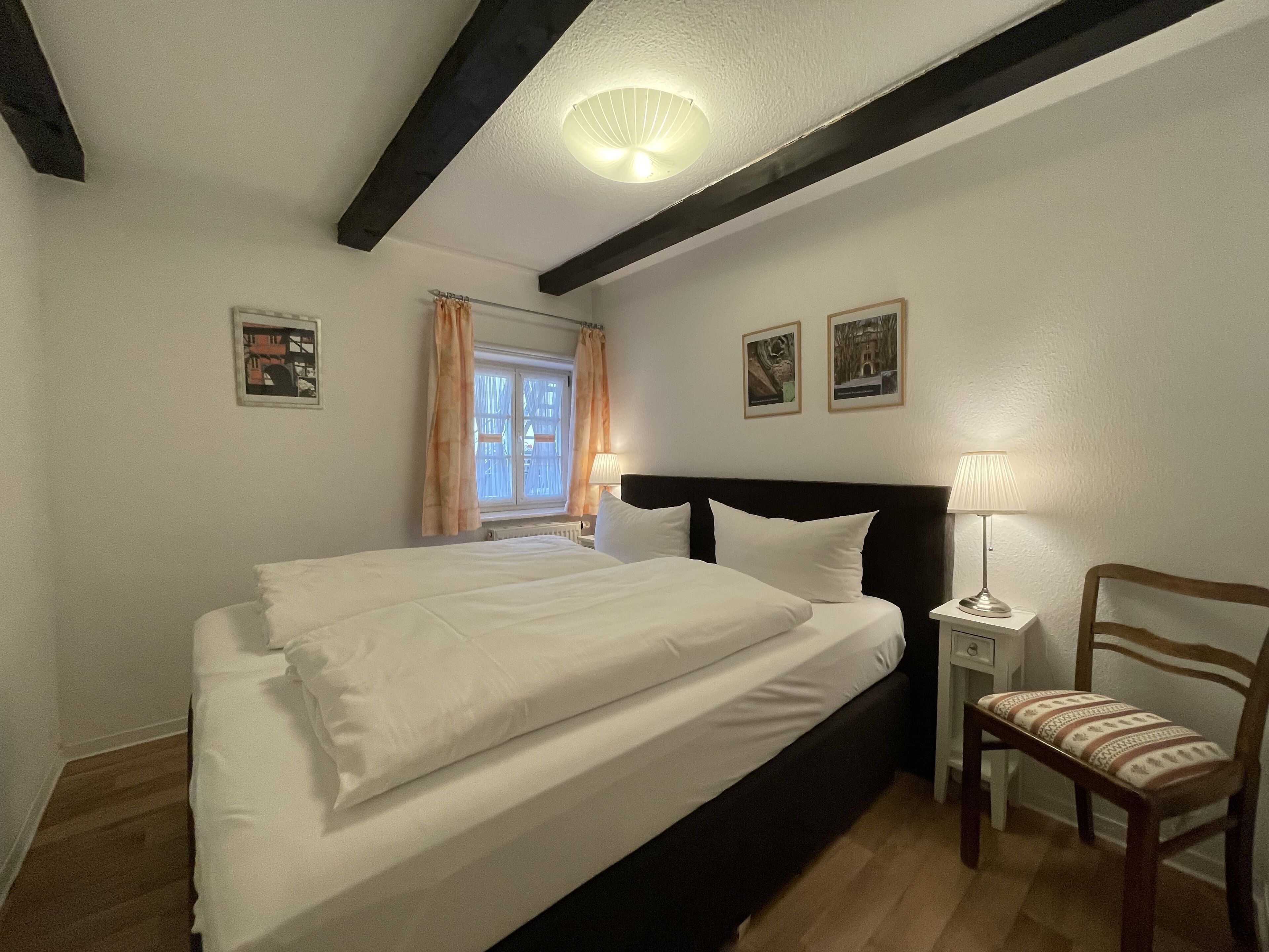 A bedroom with a large bed, a nightstand with a lamp, a chair, and two framed pictures on the wall.