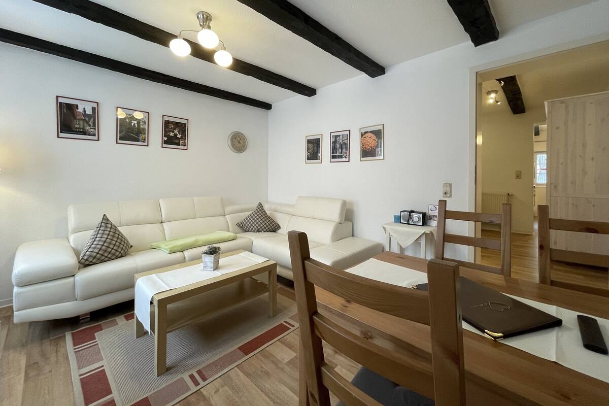 A living room with a white sofa, a wooden coffee table, and a wooden chair. There are framed pictures on the wall and a ceiling light fixture.