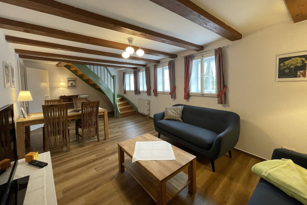 A cozy living room with a wooden floor, a dark blue sofa, a wooden coffee table, and a staircase with wooden beams.