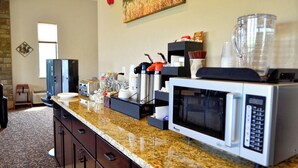 Una cocina con encimeras de granito, microondas, cafetera y licuadora. Hay una ventana con persianas y un cuadro en la pared.