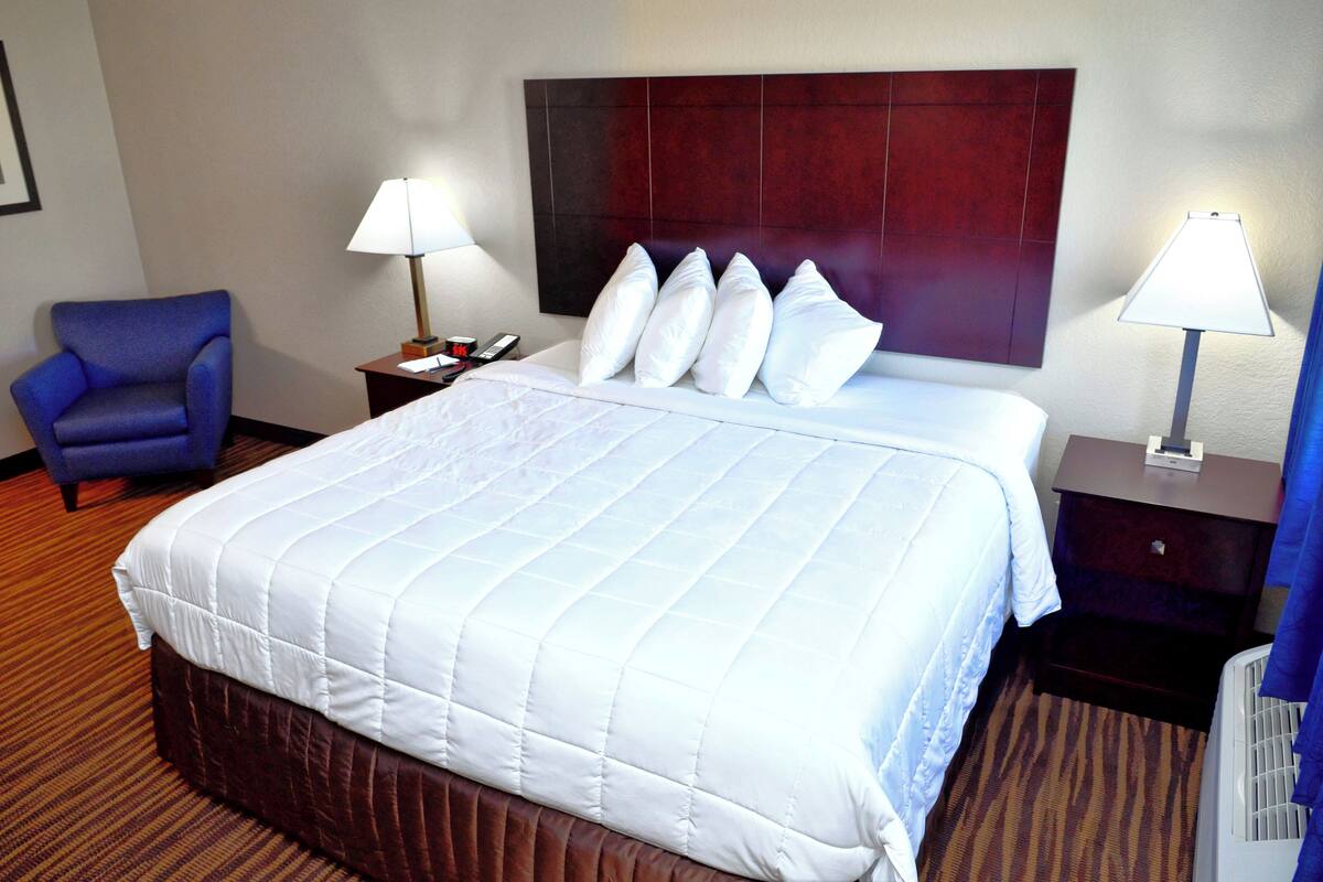 A hotel room with a large bed, two bedside tables, a blue armchair, and a wall-mounted headboard.