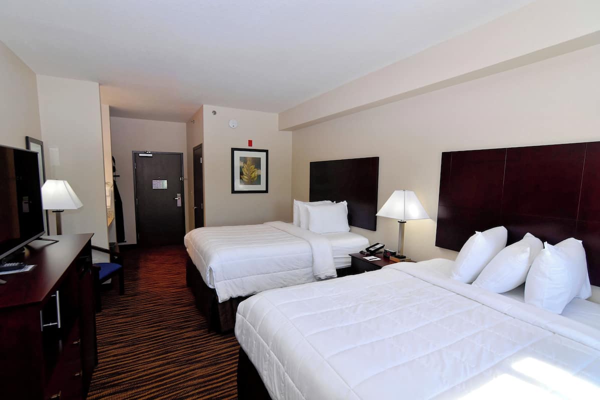 A hotel room with two beds, a desk with a lamp, a TV, and a door leading to another room.