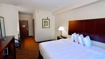 A hotel room with a large bed, a desk, a chair, a nightstand with a lamp, and a framed picture on the wall.