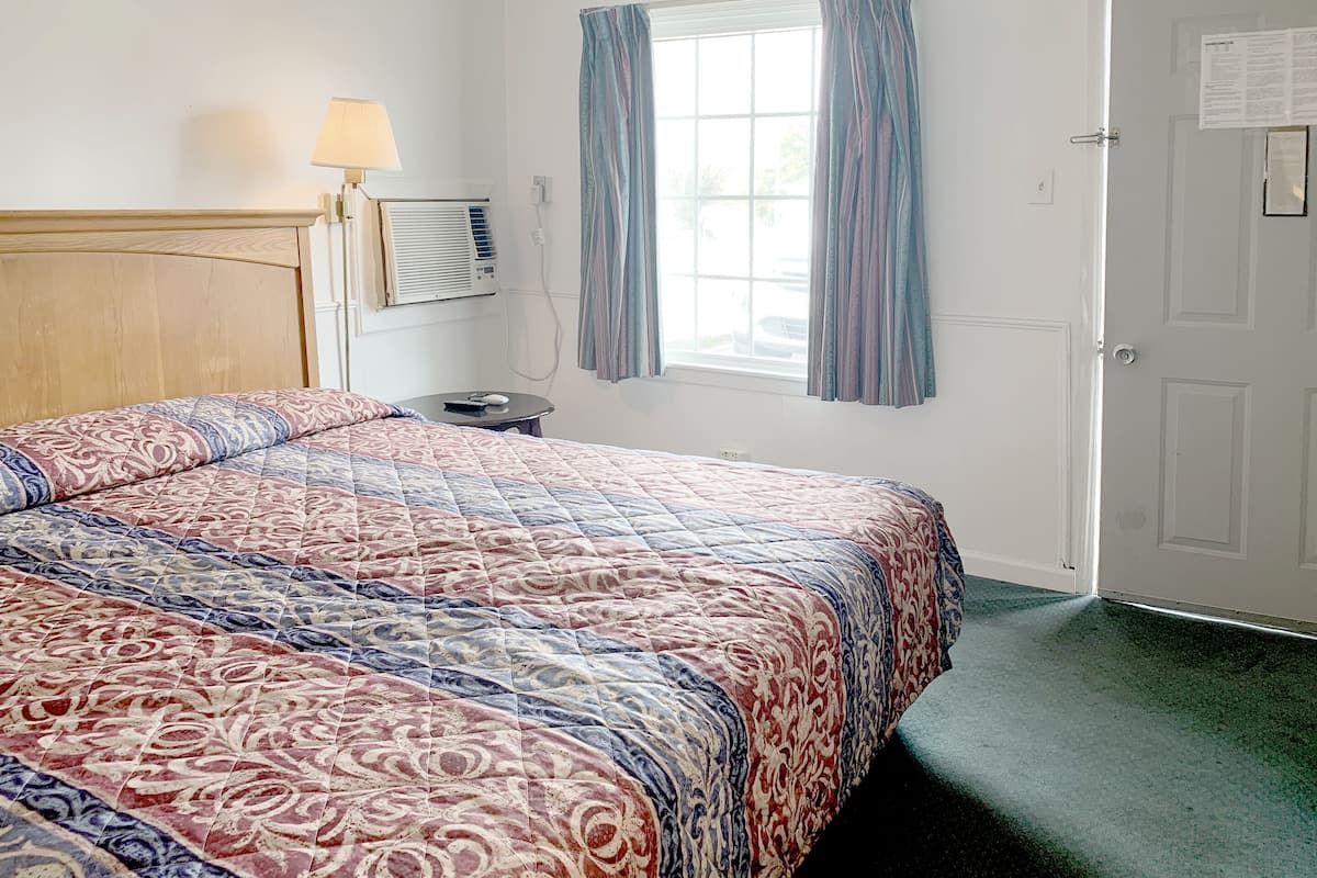 A hotel room with a bed, a bedside table with a lamp, a window with curtains, and a door.