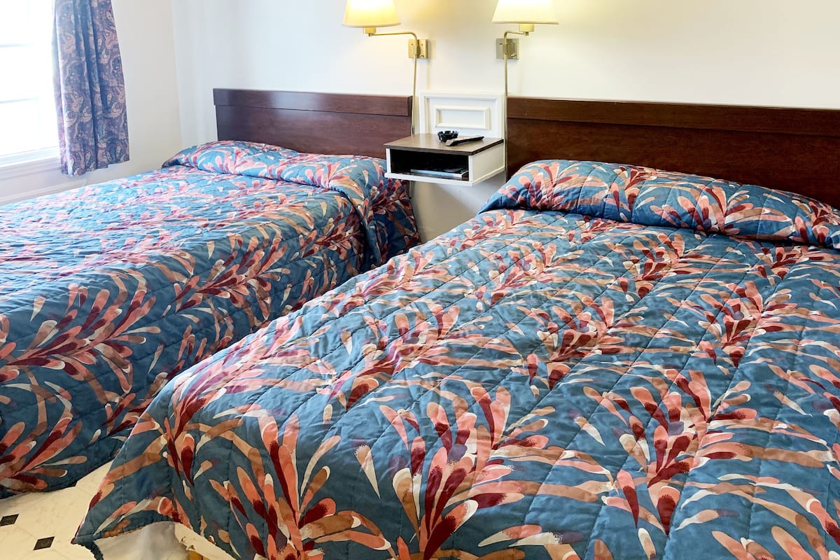 A hotel room with two beds, each with a floral-patterned bedspread. There is a nightstand with a lamp and a small shelf.
