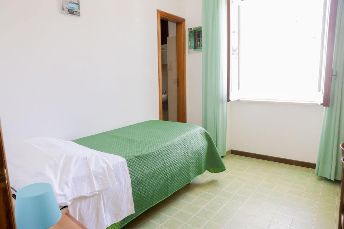 Una habitación pequeña y limpia, con una cama individual, una puerta de madera y una ventana con cortinas.