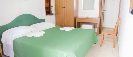A neatly made bed with white linens and a green bedspread, a wooden wardrobe, a small desk with a mirror, and a wooden chair.