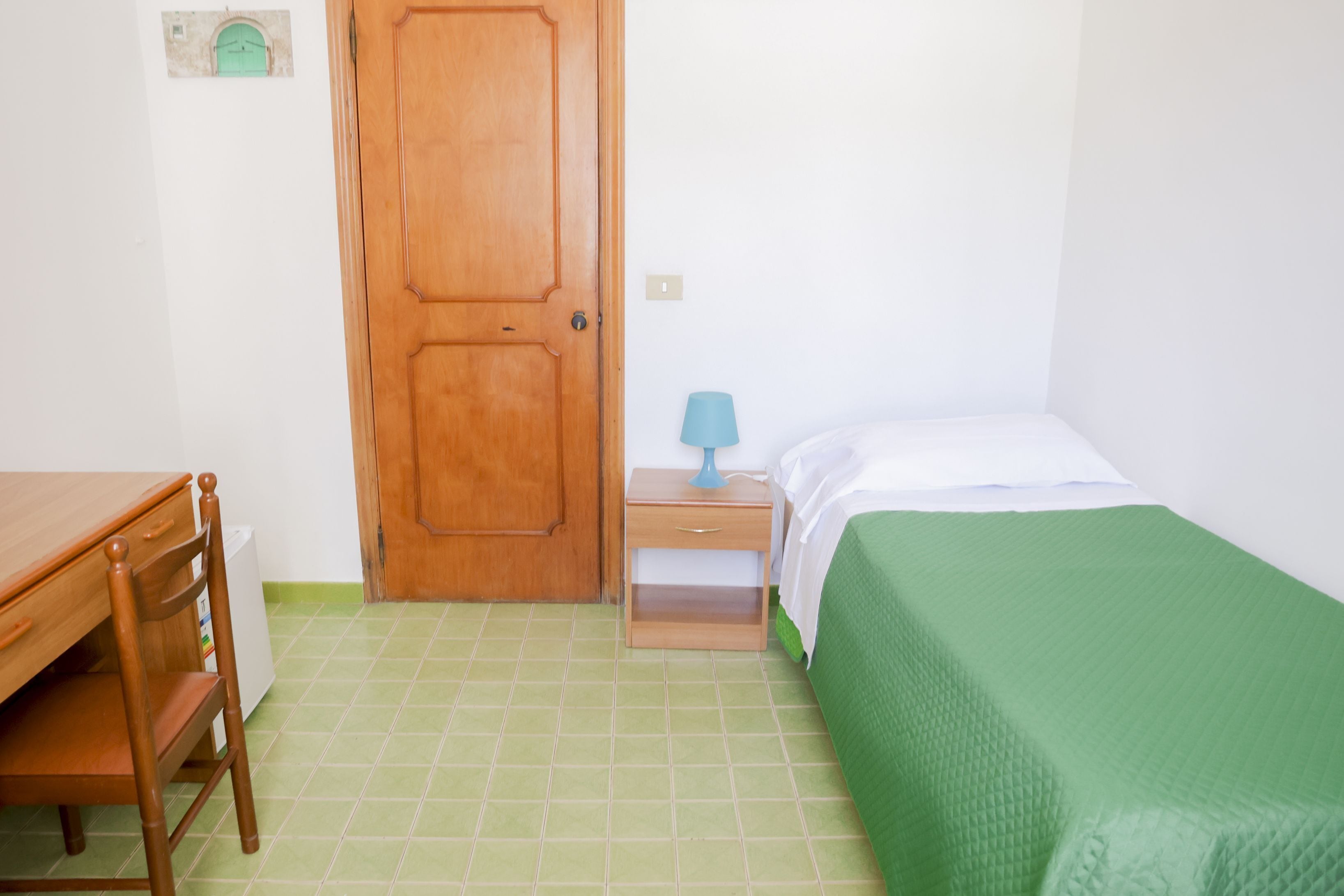 Quarto com porta de madeira, pequena mesa de cabeceira com luminária, cama com colcha verde, escrivaninha de madeira com cadeira e chão de azulejo.