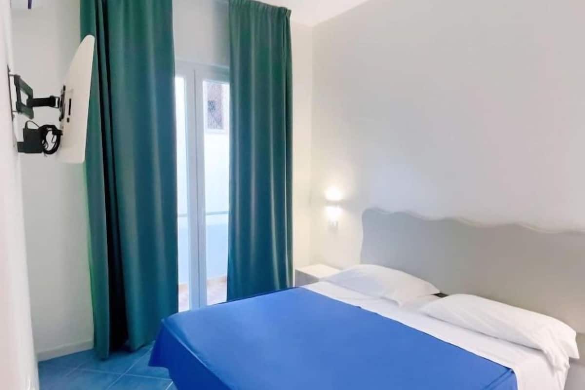 Una habitación de hotel con cama, mesita de noche, lámpara de pared y una puerta corredera con cortinas.