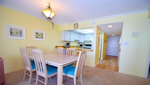 A dining area with a table and chairs, a kitchen in the background, and a carpeted floor. - Ocean Hideaway 607 by RedAwning (Ocean City)