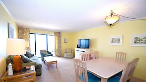 A living room with a sofa, armchair, coffee table, and a dining table with chairs. There is a television on a cabinet, and a lamp on a side table. - Ocean Hideaway 607 by RedAwning (Ocean City)