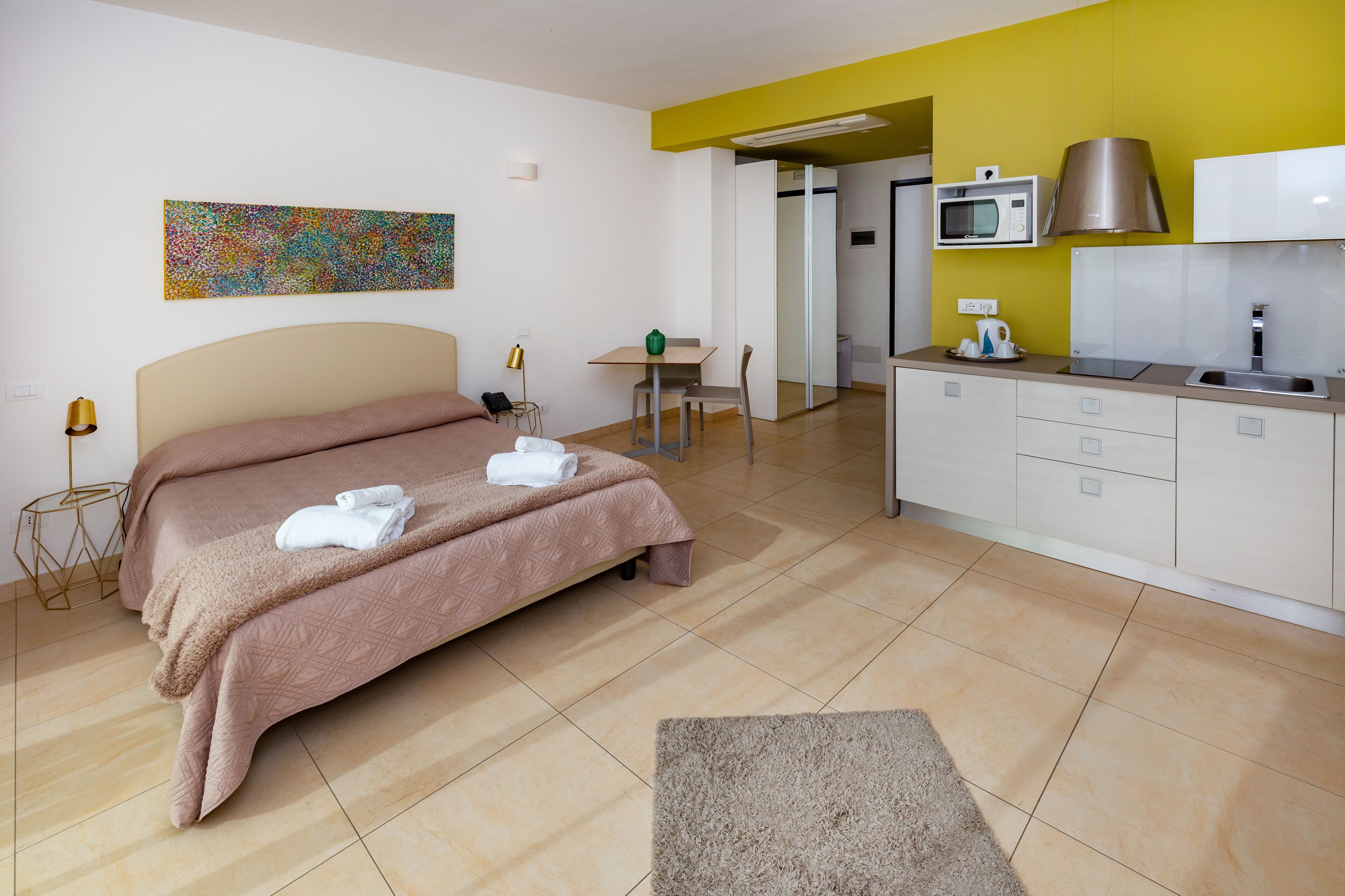 A modern hotel room with a bed, a desk, a kitchenette, and a bathroom.