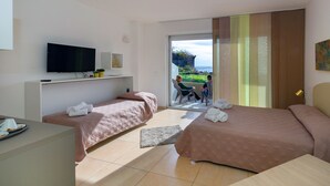 A hotel room with two beds, a TV, a desk, and a view of a balcony with people outside. - Zeus Residence Hotel (Aci Castello)