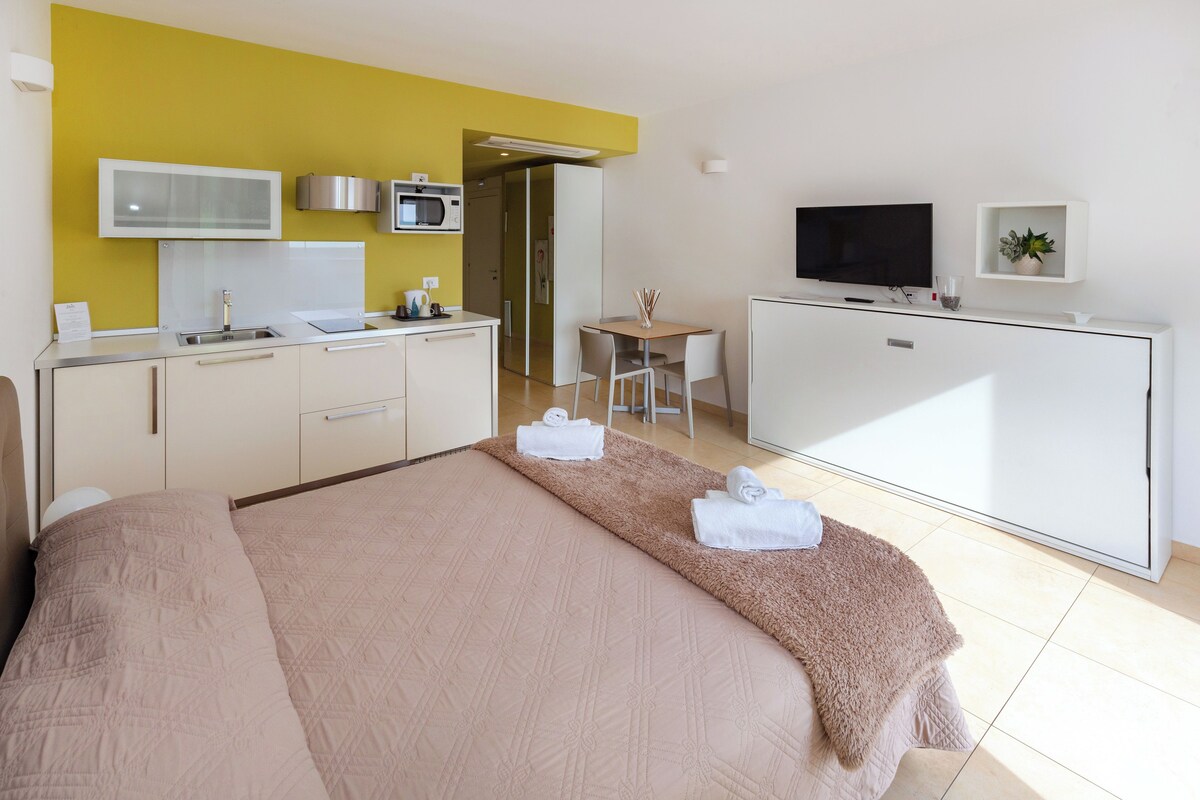 Una habitación de hotel con cama, kitchenette, televisor y una pequeña mesa.