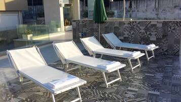 Un área de lounge en la azotea con cuatro tumbonas blancas, barandilla de vidrio y pared con azulejos estampados.