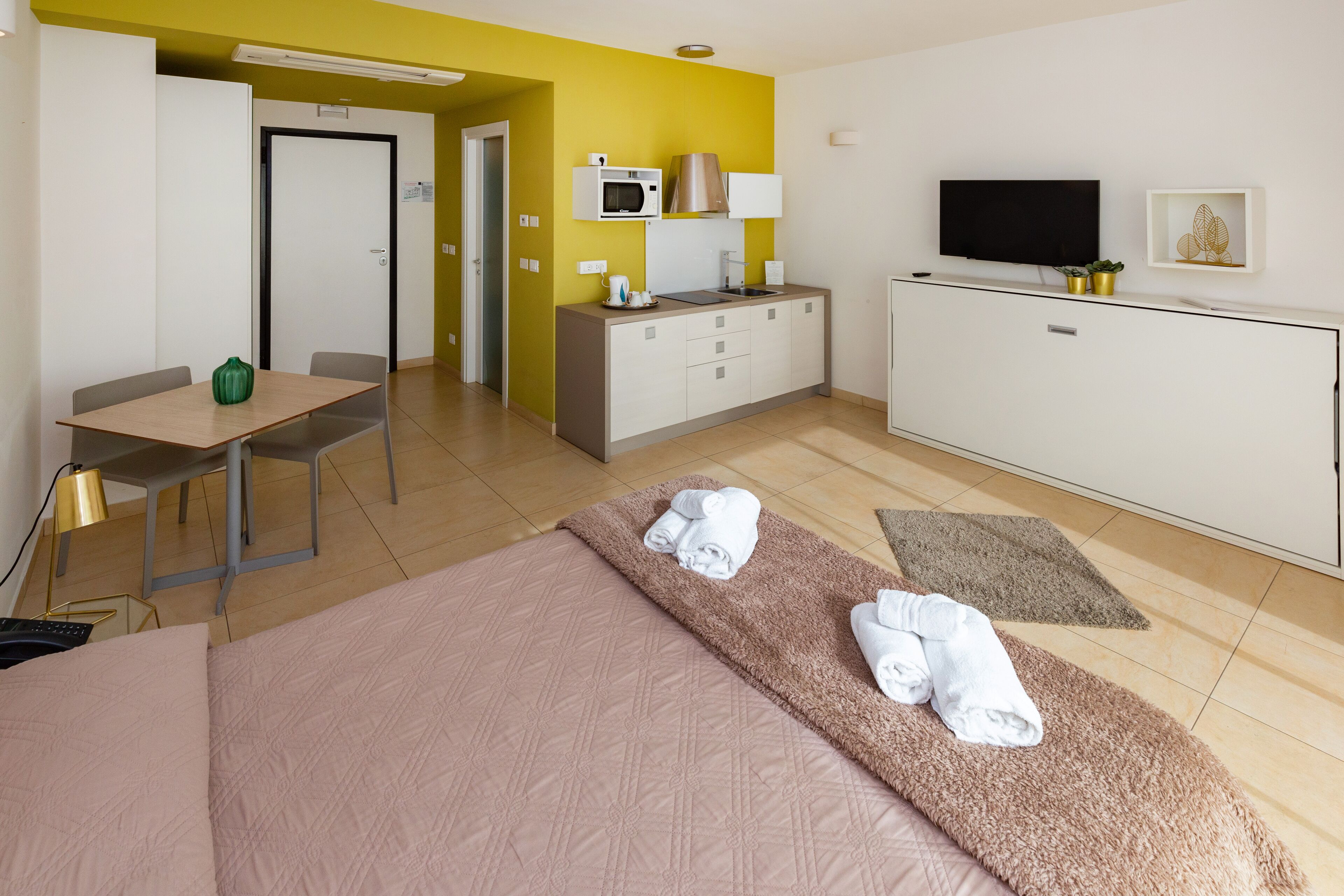 A hotel room with a bed, a desk, a chair, a kitchenette, and a TV.