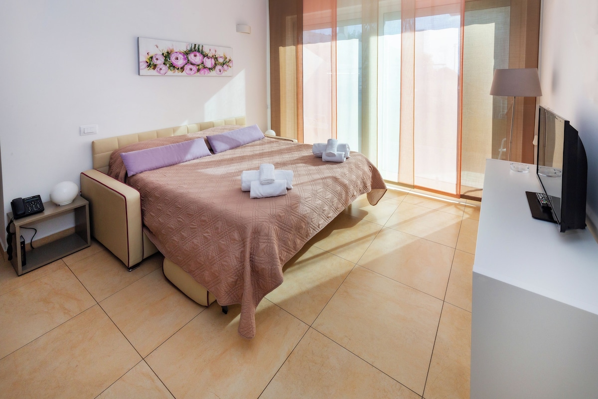 A modern hotel room with a large bed, a bedside table with a lamp, a TV, and a view of the outside through a large window.