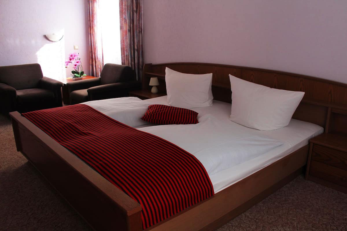 A neatly made bed with white linens and a red striped blanket, a wooden headboard, a small bedside table with a lamp, and a chair with a table beside the window.