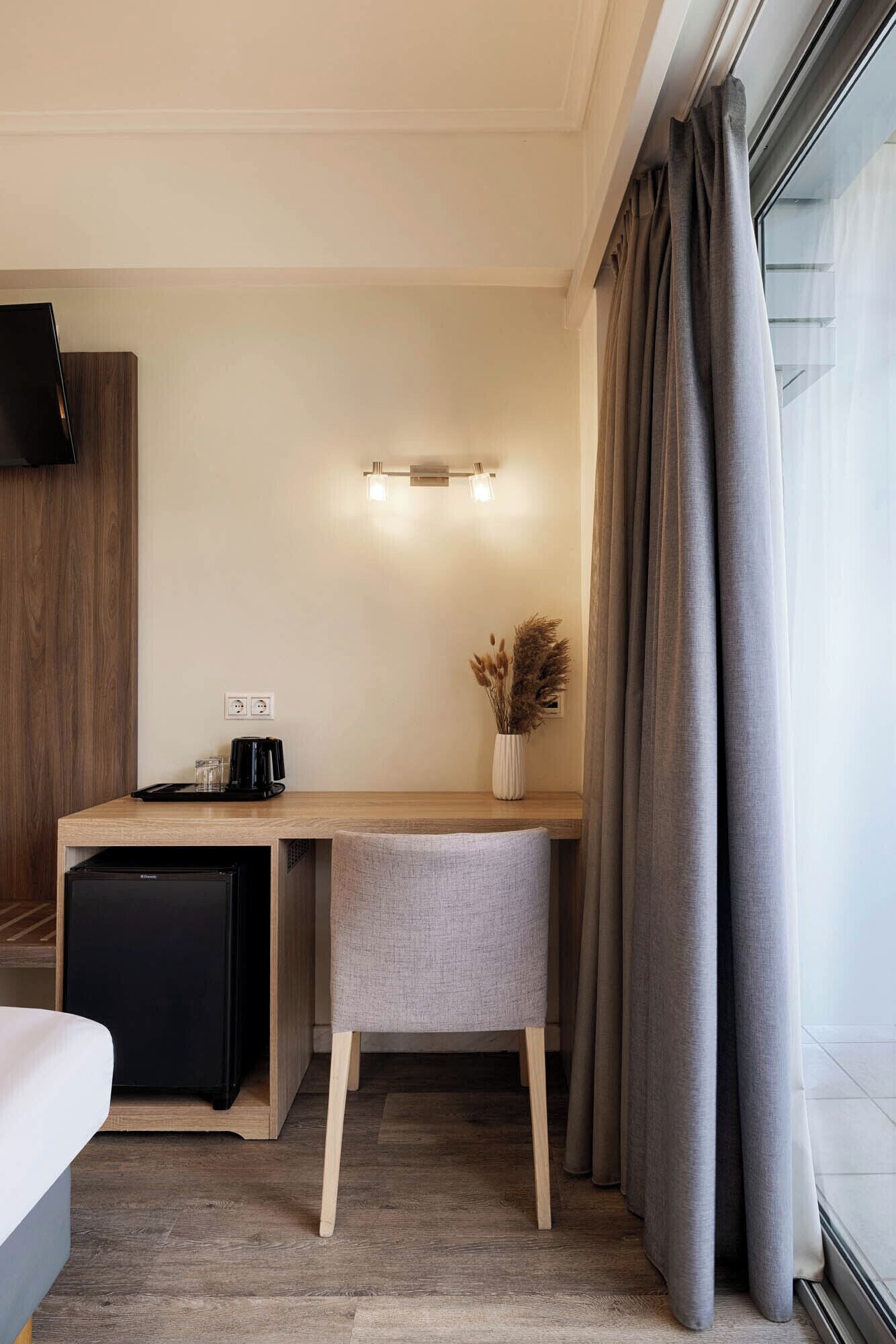 Habitación de hotel con un escritorio de madera, una kitchenette pequeña, una silla blanca, una mesita de noche y una ventana con cortinas.