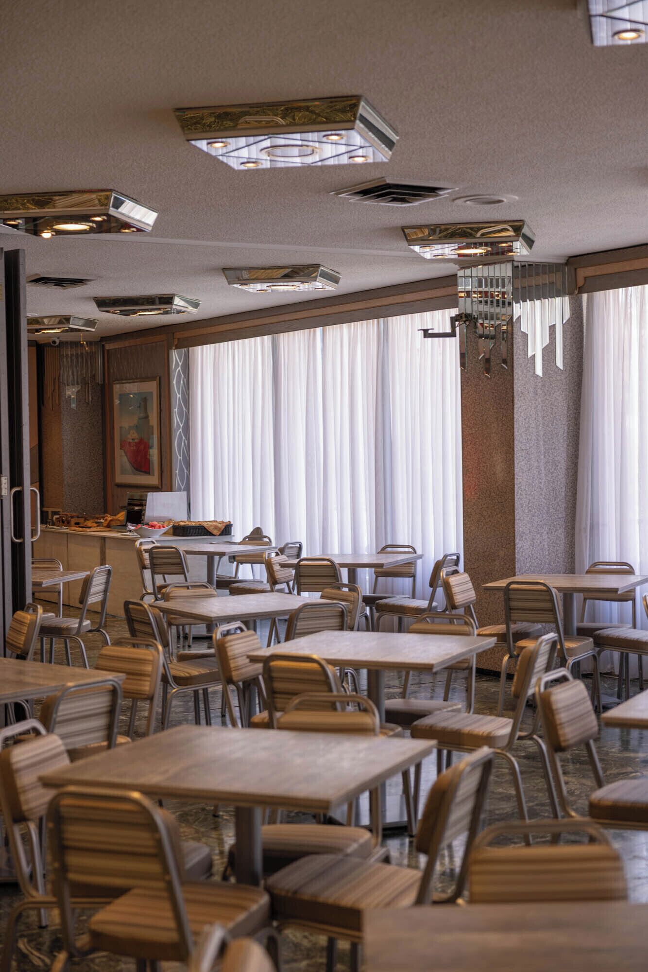 A hotel dining area with multiple tables and chairs, a counter with a coffee machine, and large windows with curtains.