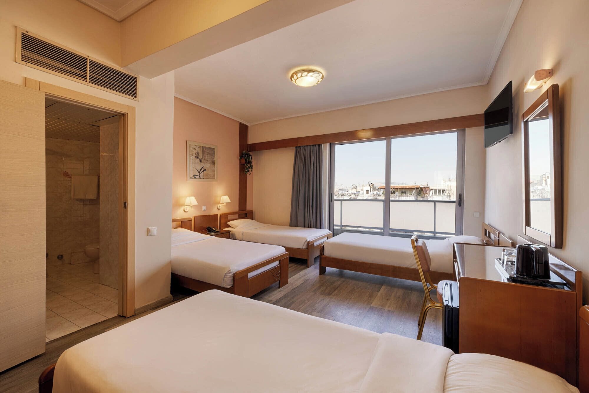 A hotel room with two beds, a desk, a chair, a TV, and a large window with a city view.