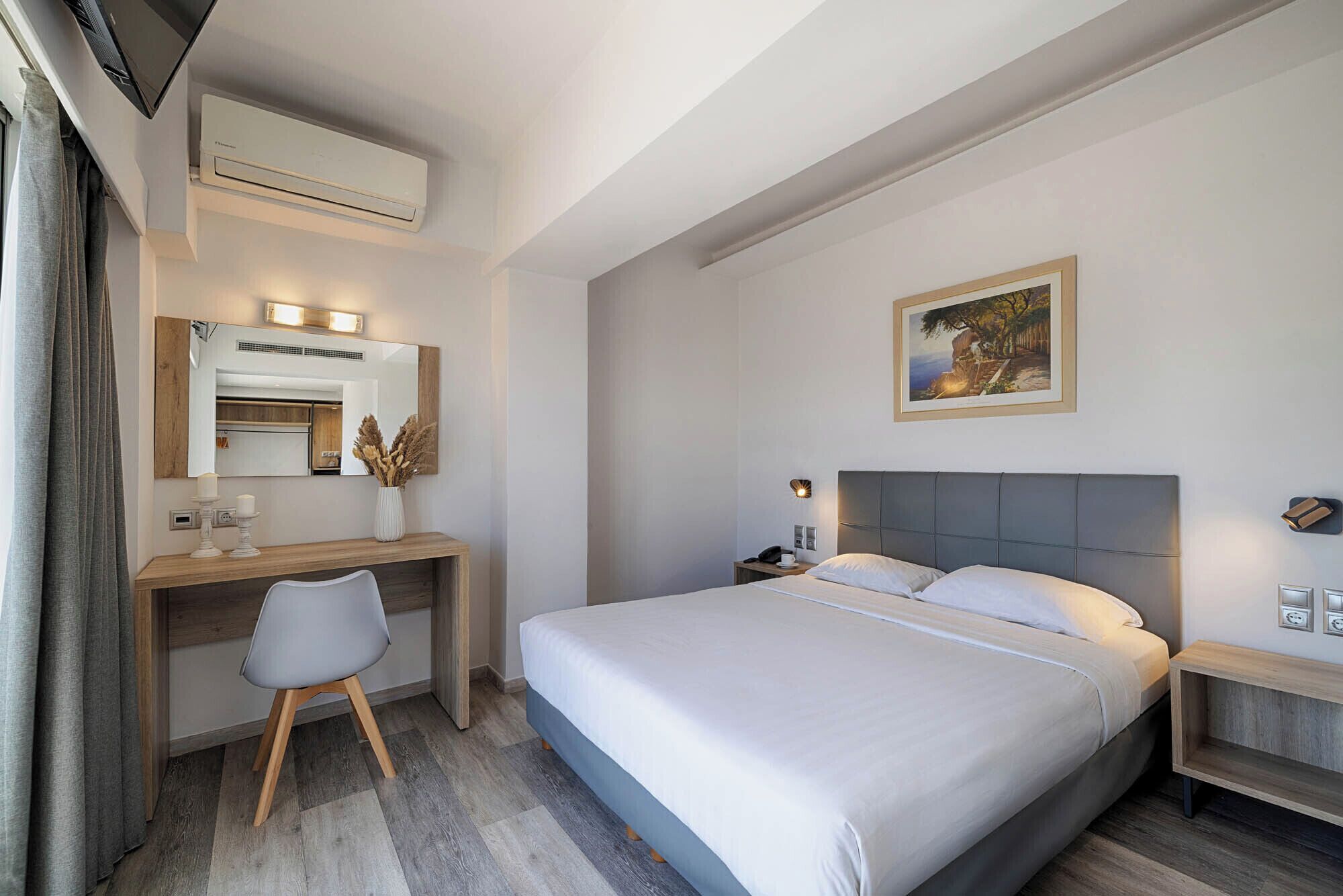 A hotel room with a bed, bedside tables, a desk with a chair, a mirror, and an air conditioning unit.