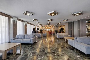 Um hall de hotel com piso de mármore, sofás cinzas e luminárias no teto.