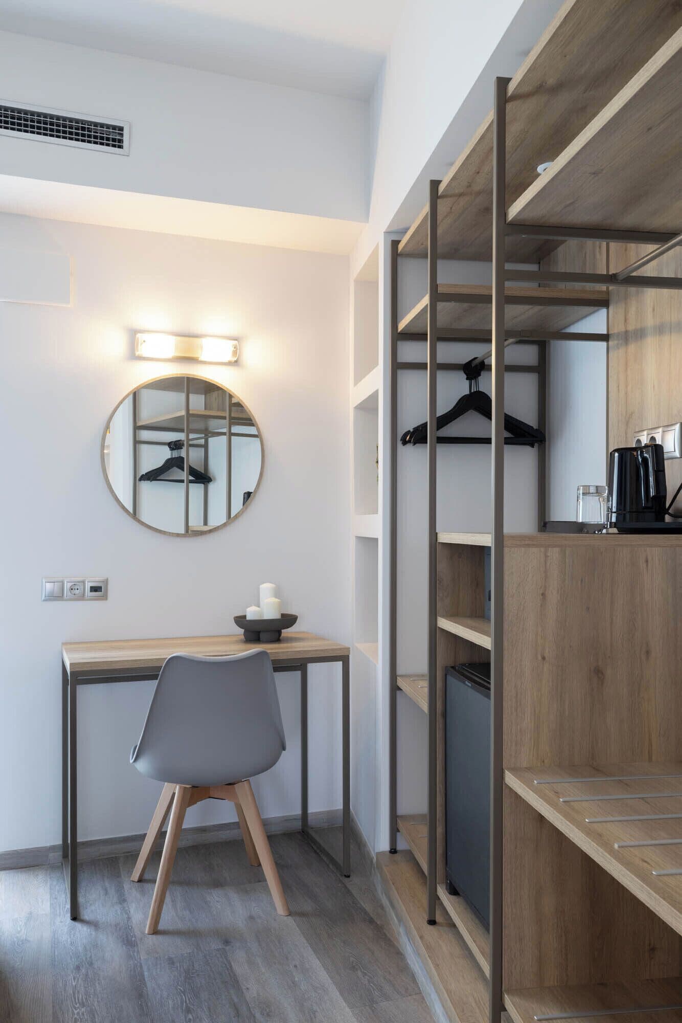Une chambre d’hôtel moderne avec un bureau en bois, une chaise, un miroir rond et une armoire intégrée avec un espace pour suspendre les vêtements.