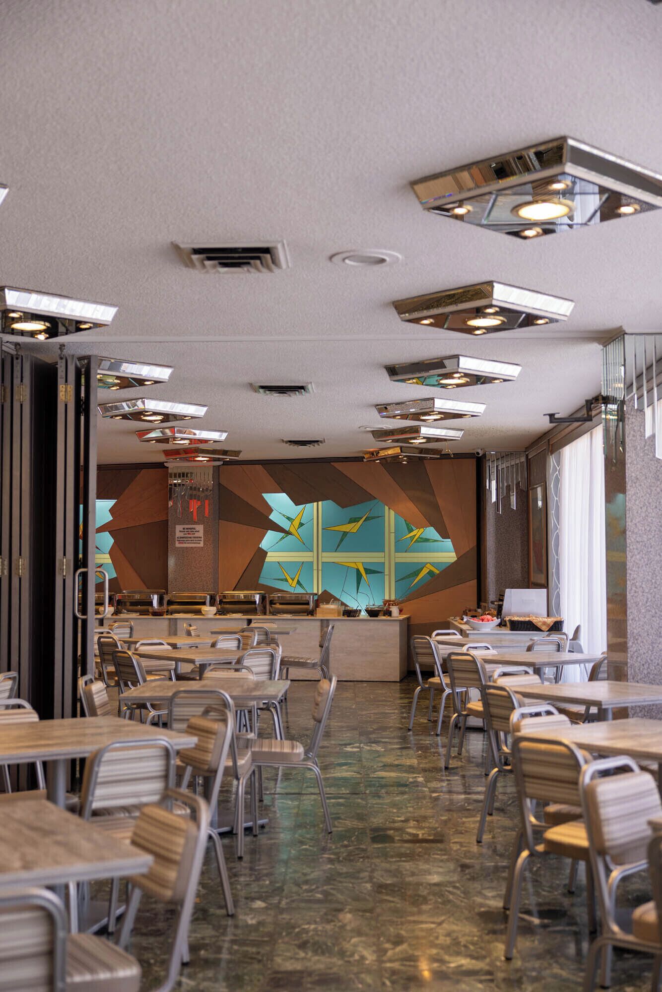 A modern dining area with multiple tables and chairs, a decorative wall with geometric patterns, and a ceiling with recessed lighting.