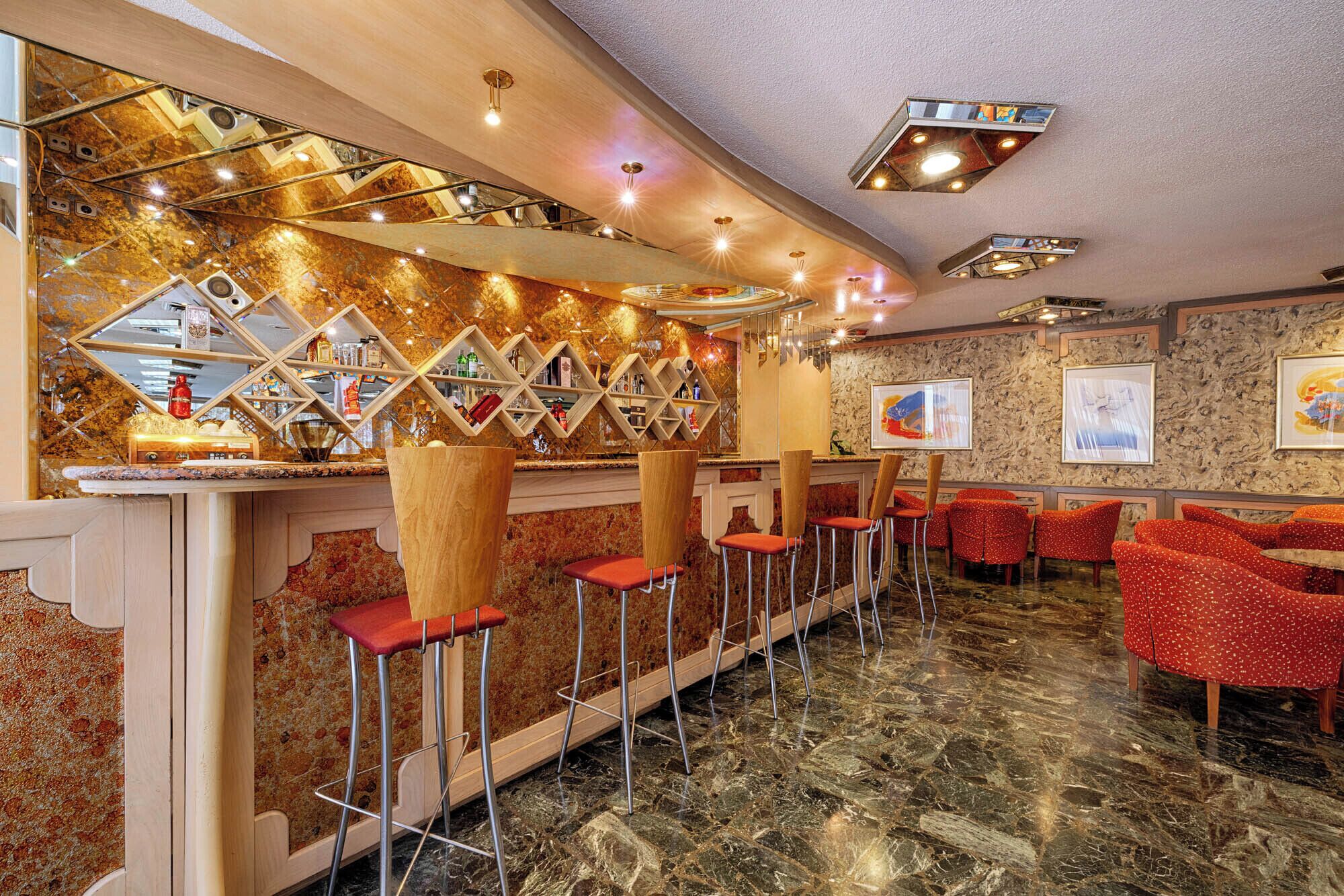 A bar area with a marble floor, red bar stools, and a mirrored wall with shelves of bottles.