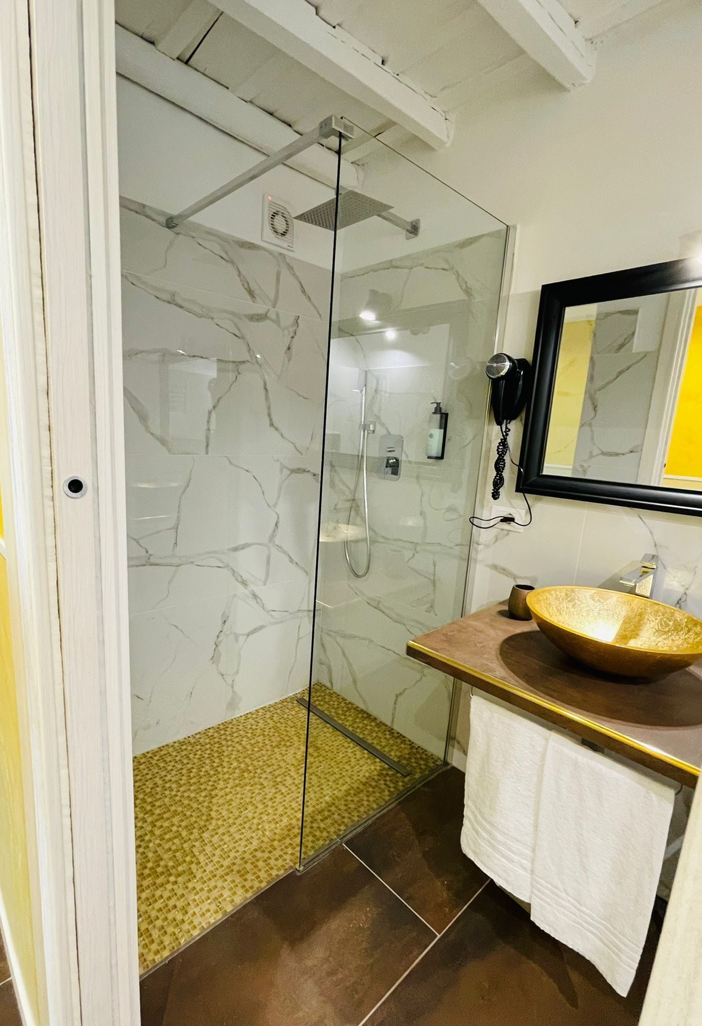 Un baño moderno con cabina de ducha de vidrio, pared revestida de mármol, tocador de madera con lavamanos grande y un espejo sobre el lavamanos.