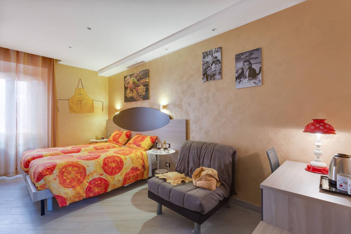 A hotel room with a bed, a sofa, a desk, and a chair. There are wall art pieces and a lamp on the desk.